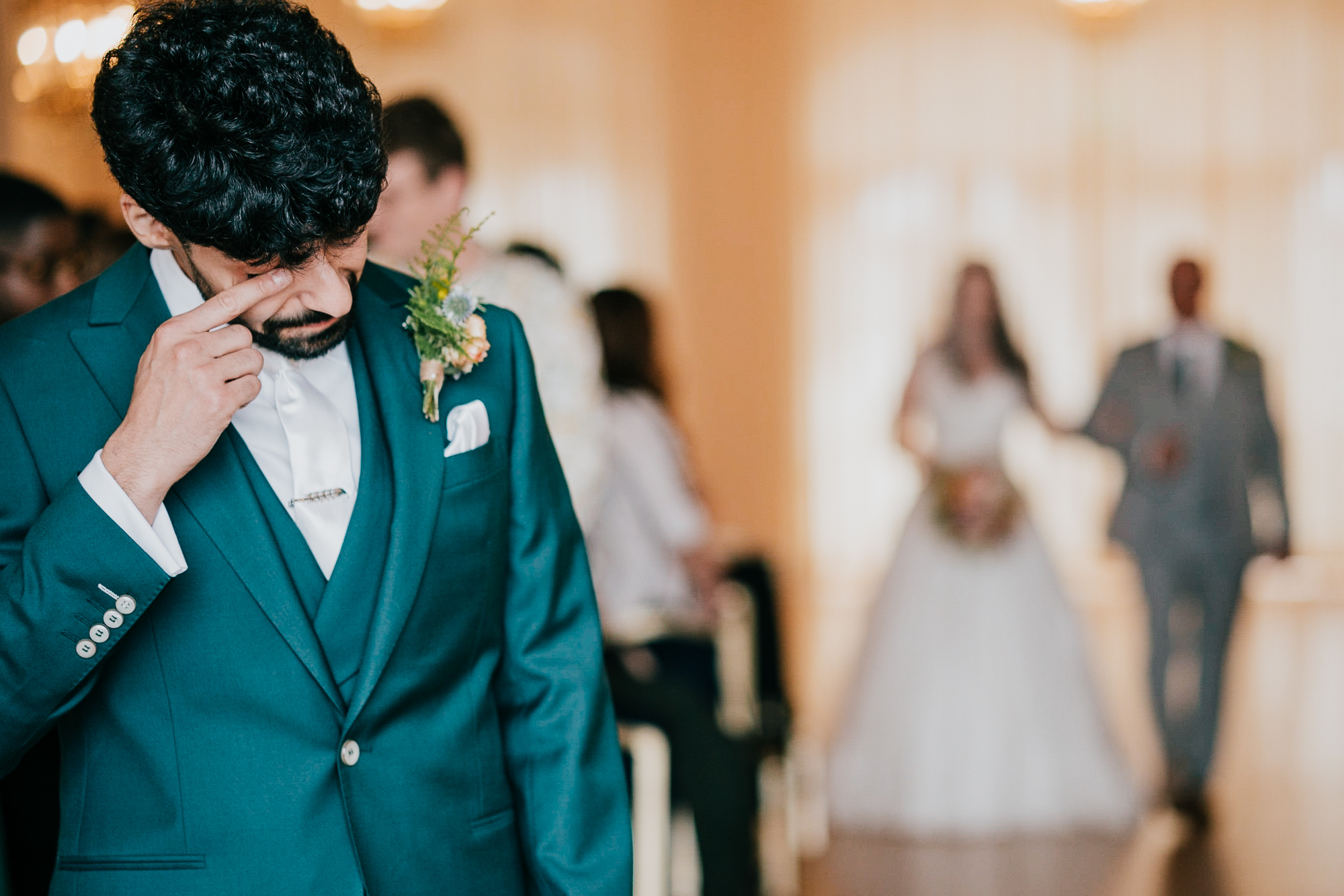 Groom wipes away a tear as his bride, slightly out of focus, walks down the aisle to meet him, at their wedding ceremony at The Atlantic Hotel, Newquay.