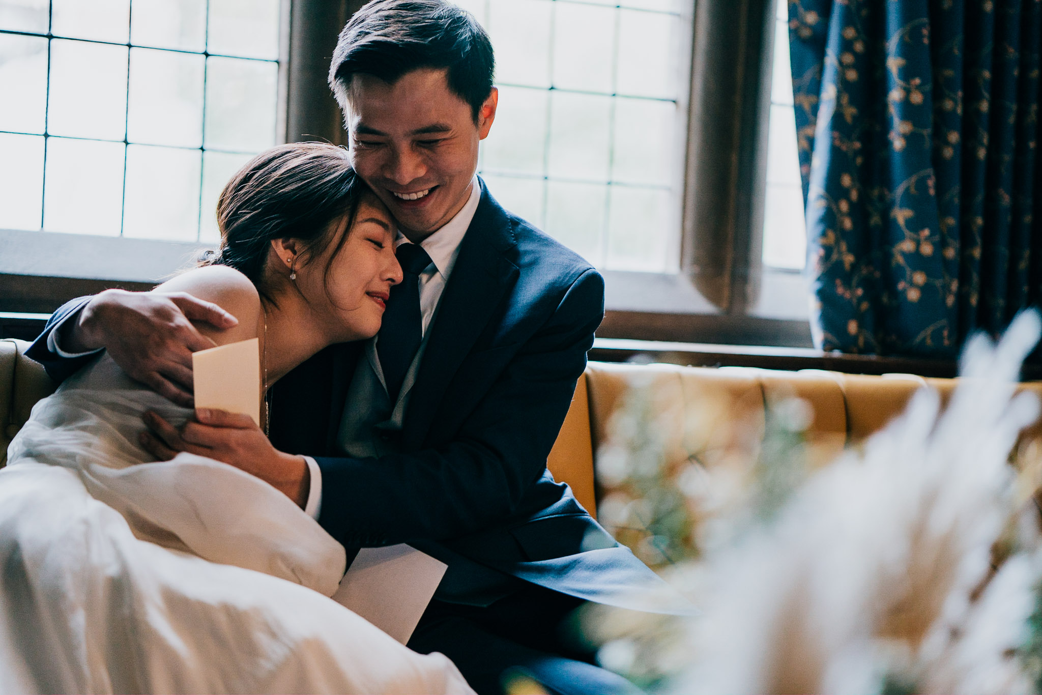 Bride and groom hug on a sofa indoors, smiling as warm daylight pours in from a large window behind them.