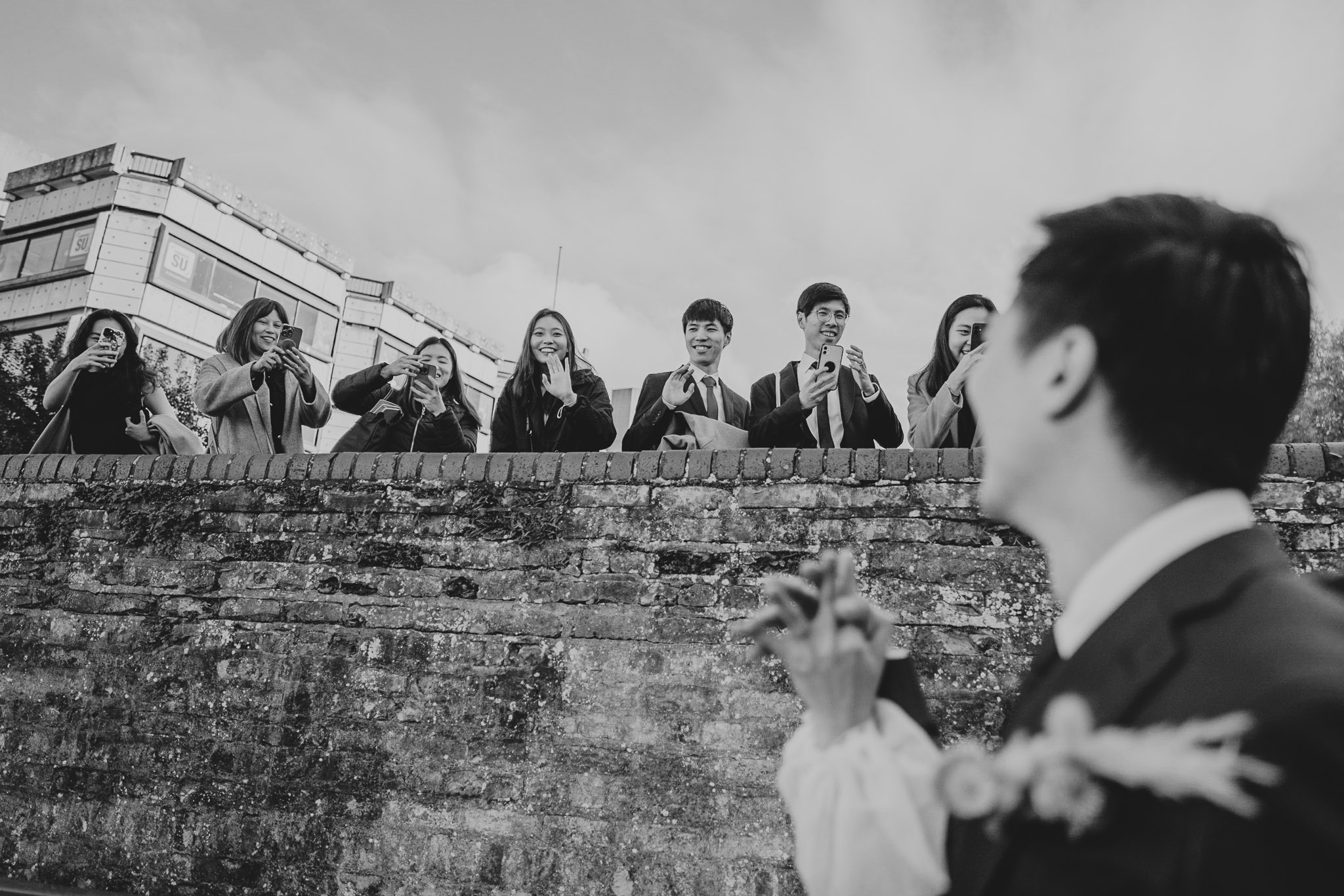 Group of people in business attire standing behind a brick wall, taking photos with smartphones; foreground person in suit is out of focus.