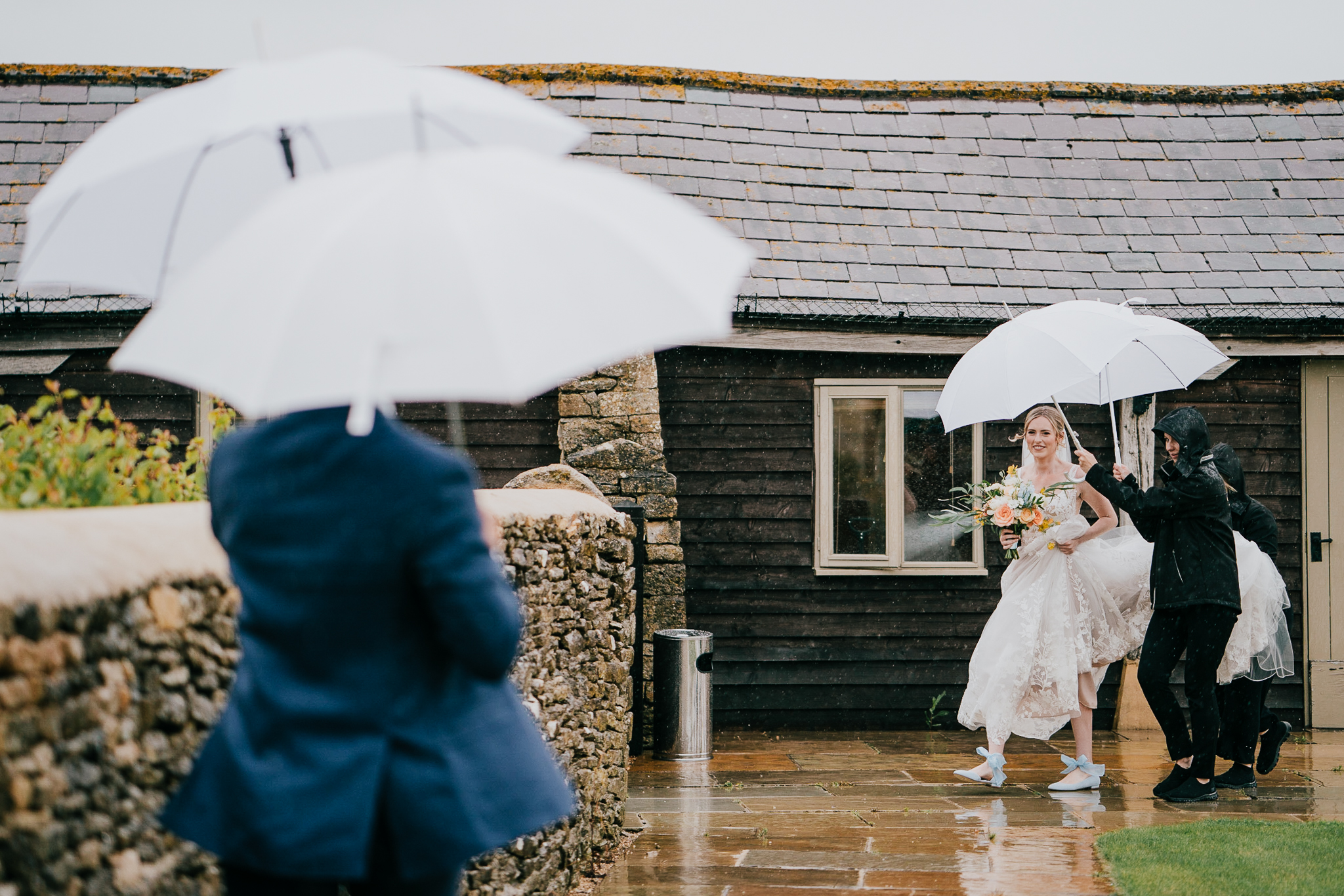 The bride battles the rain, with people trying to protect her with umbrellas, on her way to the wedding ceremony at Caswell House.