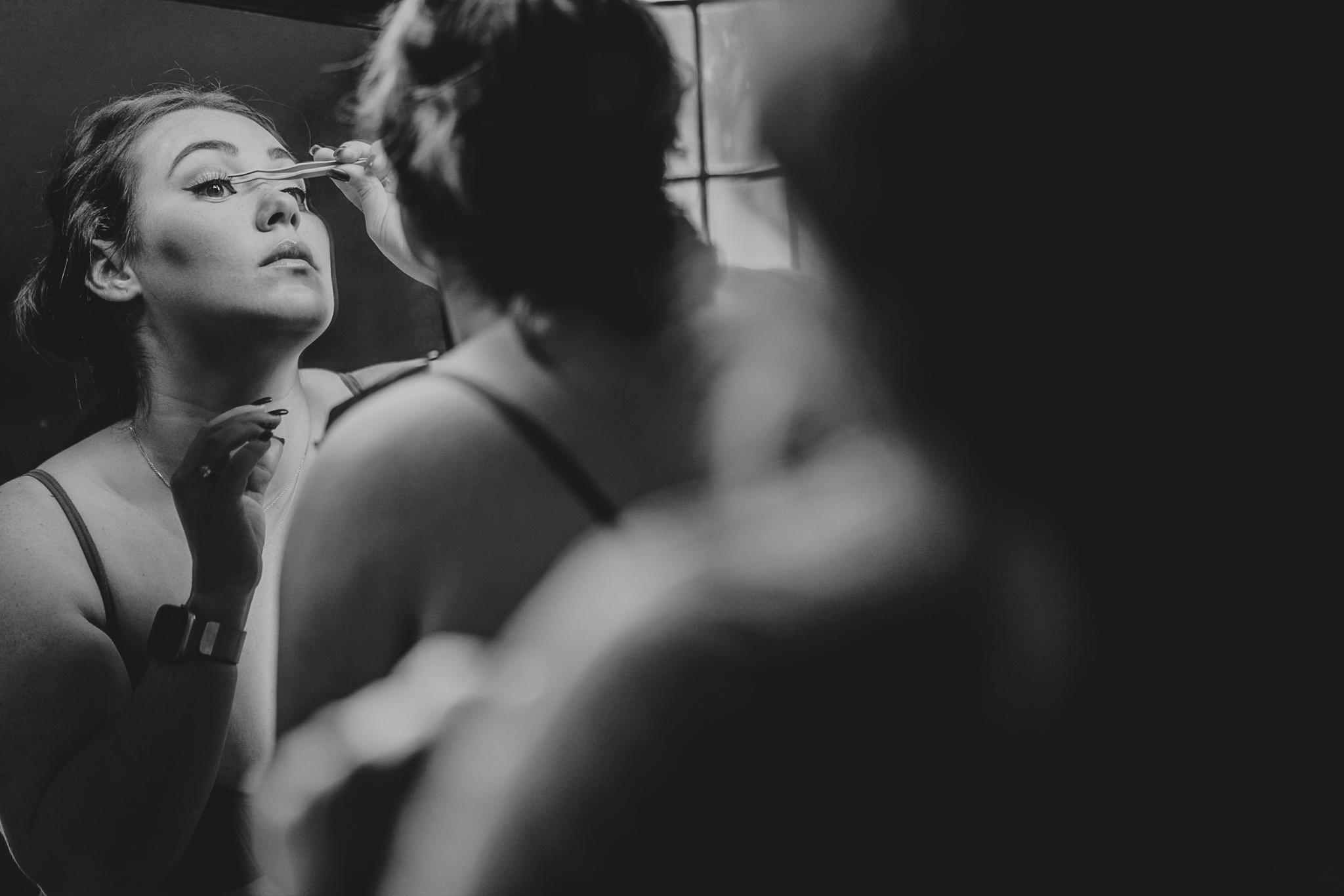 pengersick-castle-wedding-photographer 1 – Alan Law Photography A bridesmaid applying makeup, viewed over the shoulder of a different bridesmaid, and through a mirror.