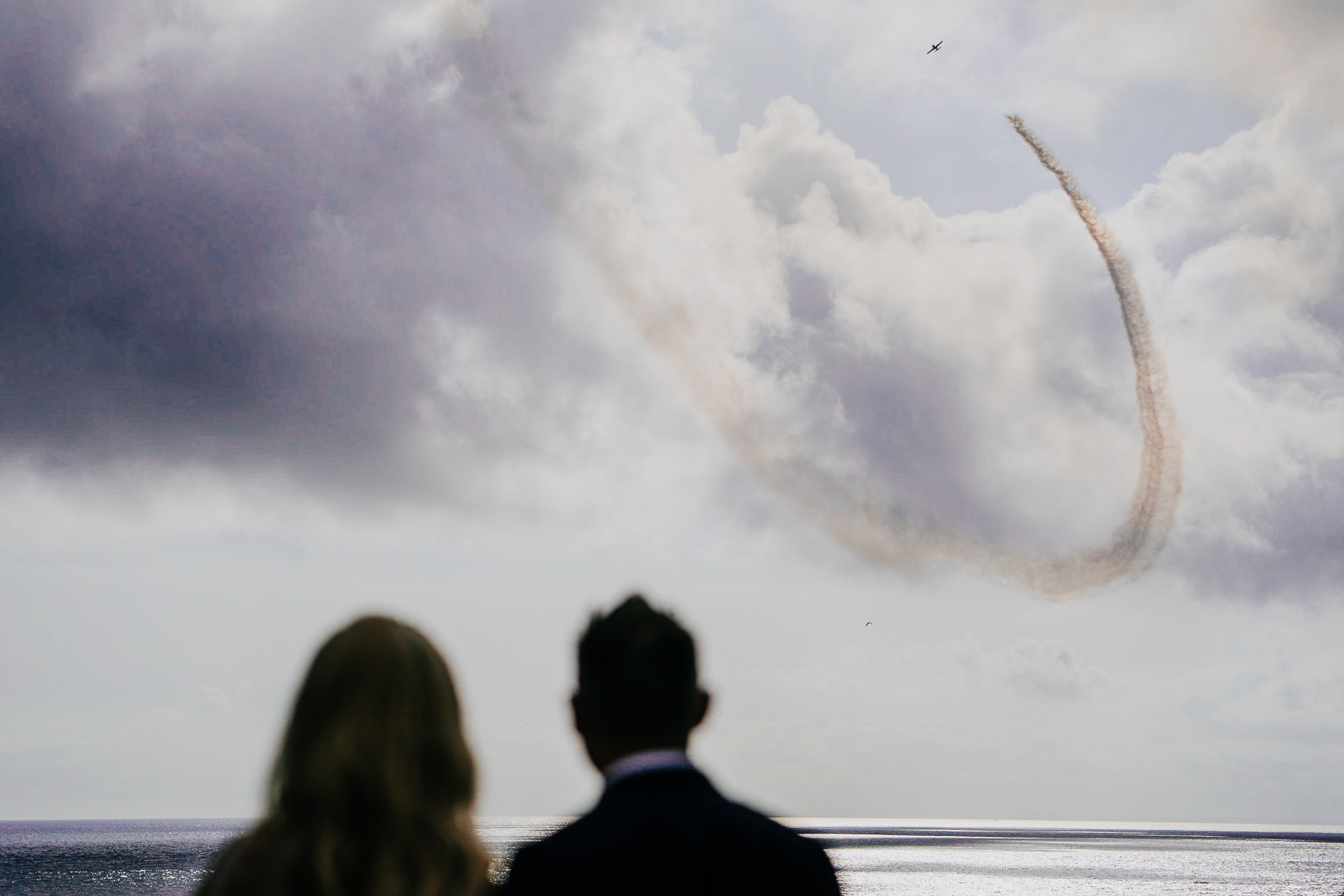 A stunt plane flies overhead and leaves a smoke trail, as the bride and groom watch on below, at Polhawn Fort, Cornwall.