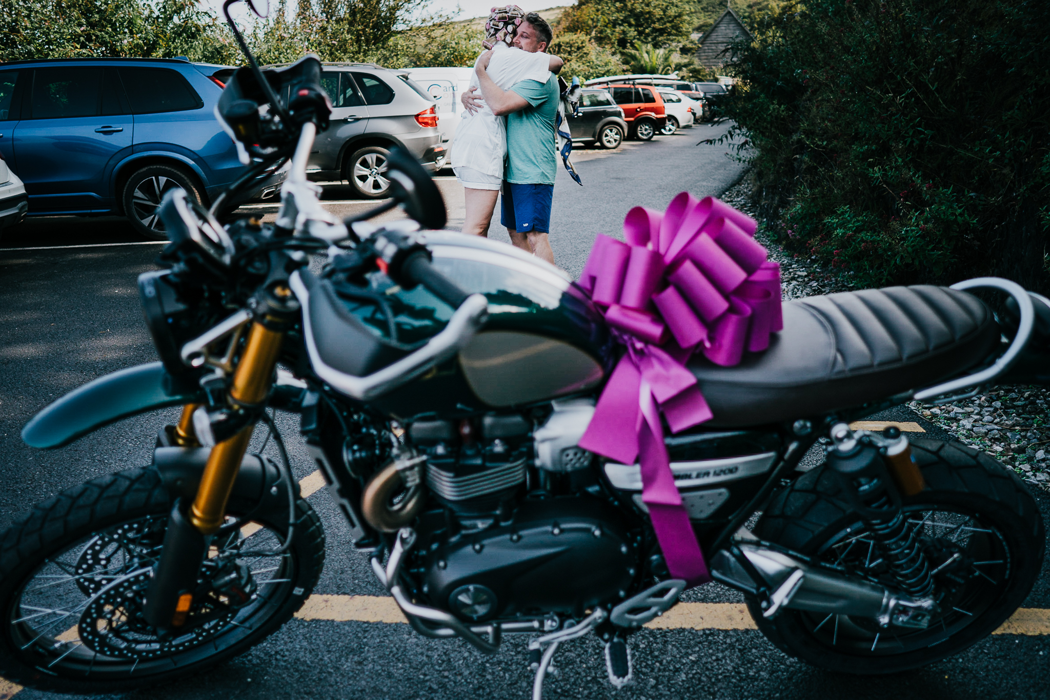 The bride and groom hug in the background; the foreground shows the motorbike, wrapped in a bow, that the bride surprised the groom with.
